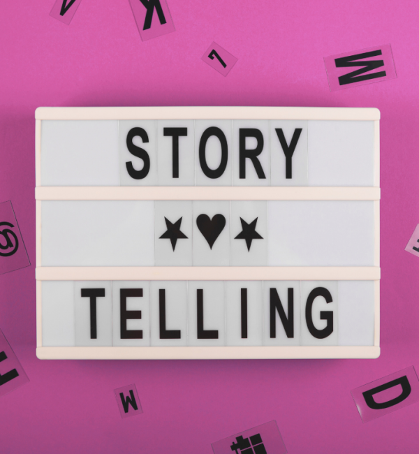 A 3 lined, white light box with the words, story on the first line, telling on the 3rd line. A heart image is on the second line. with a pink background