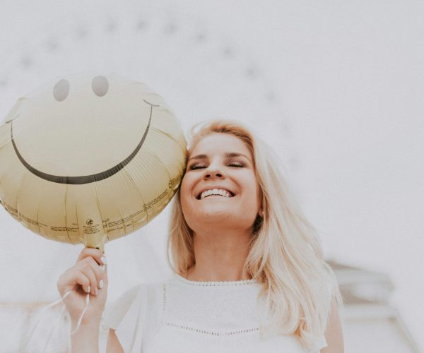 a blonde women, smiling whilst holding a smiley face balloon